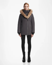 A woman faces forward against a plain white background, wearing the Rudsak AURORE - L123561 Grey fitted winter coat with a large fur-trimmed hood, paired with black leggings and black ankle boots.