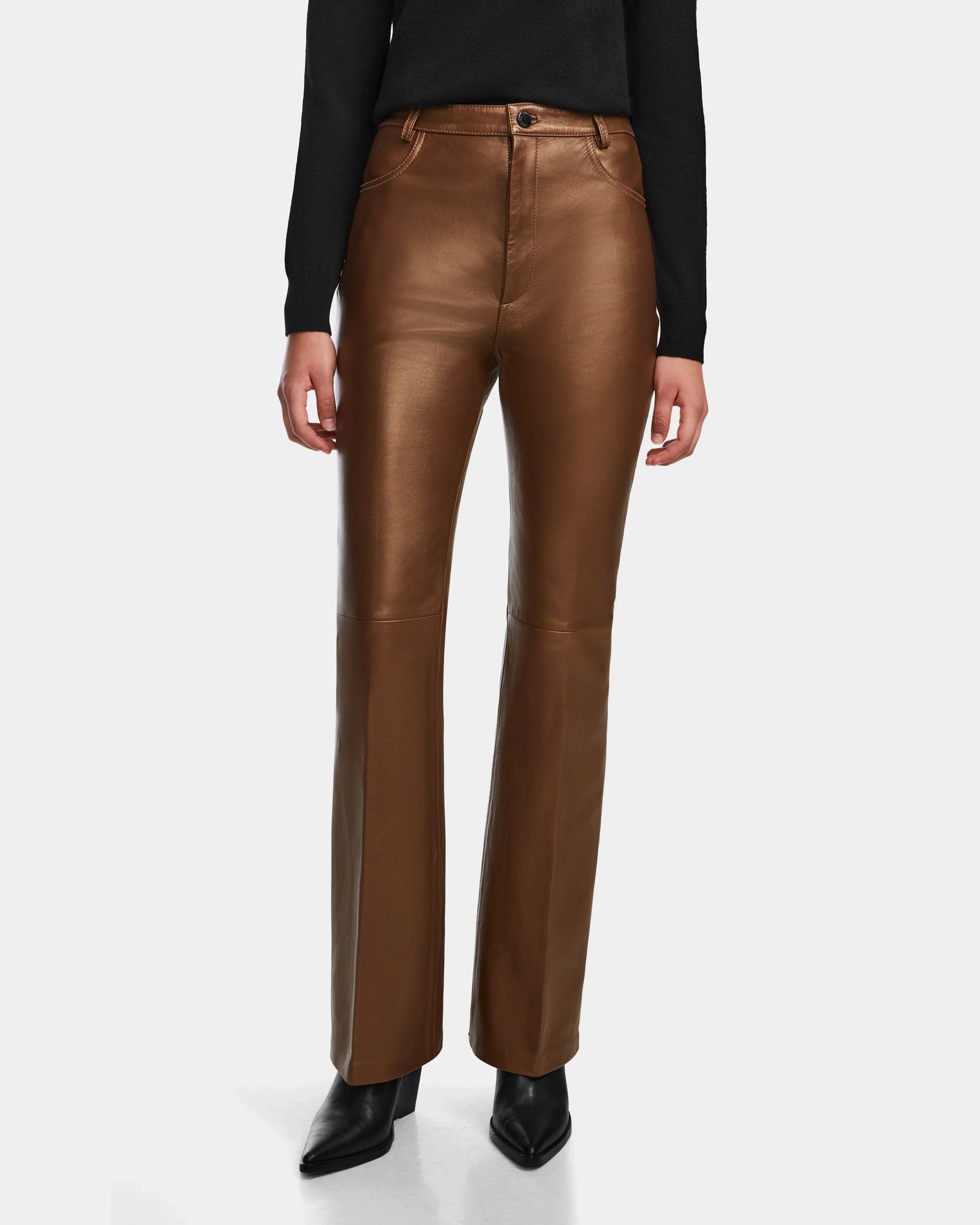 A person wears Rudsak’s CARISSA - 8524545 Antique Bronze high-waisted leather pants with a slight flare, black pointed-toe shoes, and a long-sleeve black top, standing against a plain white background.