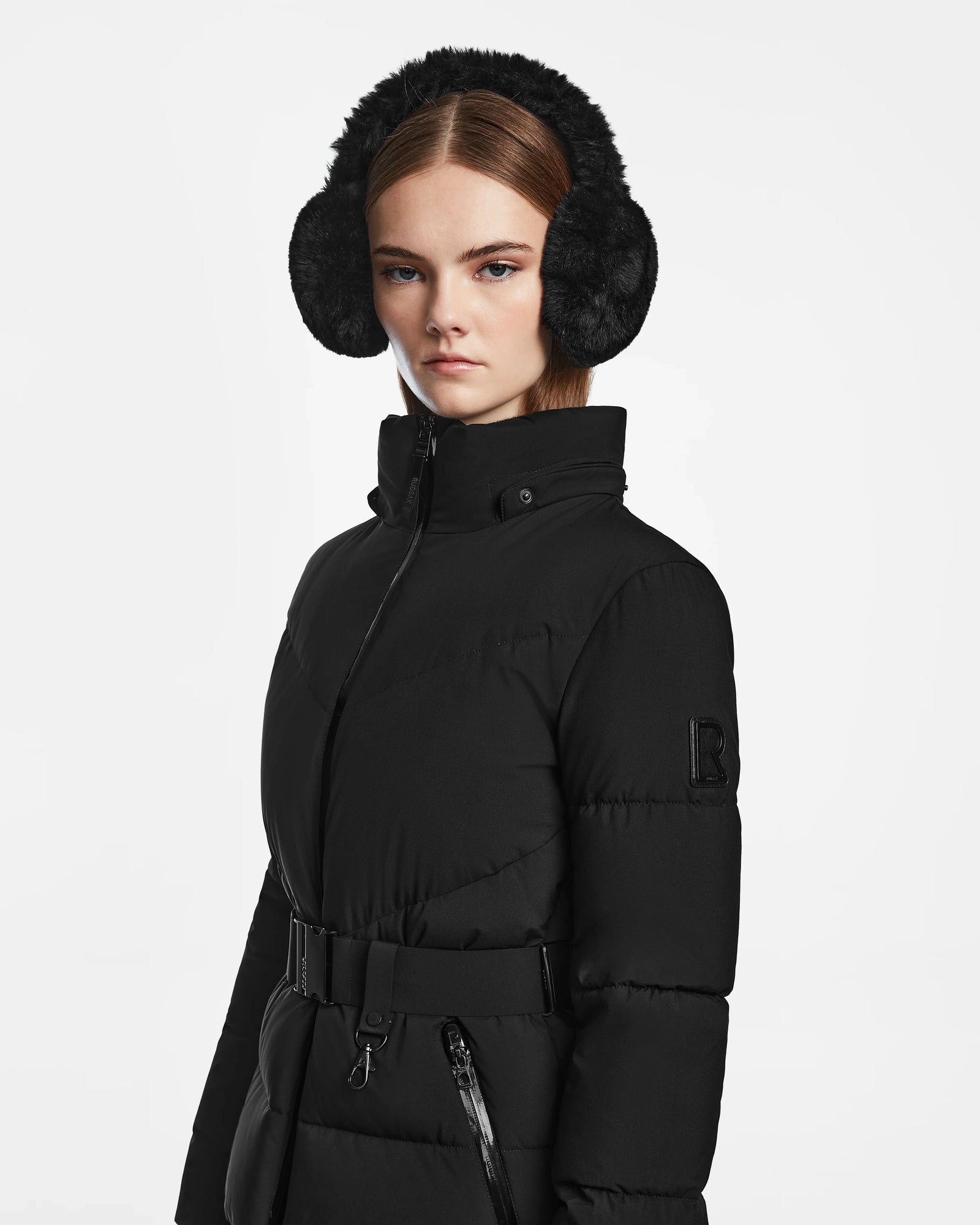 A young woman in Rudsak’s CHRISTELLE M - L123525 Black belted women's quilted down puffer with a fitted silhouette and black earmuffs stands against a white background, looking directly at the camera with a neutral expression.