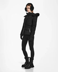 A woman with straight brown hair and a neutral expression models the Rudsak CHRISTELLE M - L123525 Black quilted down puffer, styled with earmuffs, leggings, gloves, and lace-up boots against a white background.
