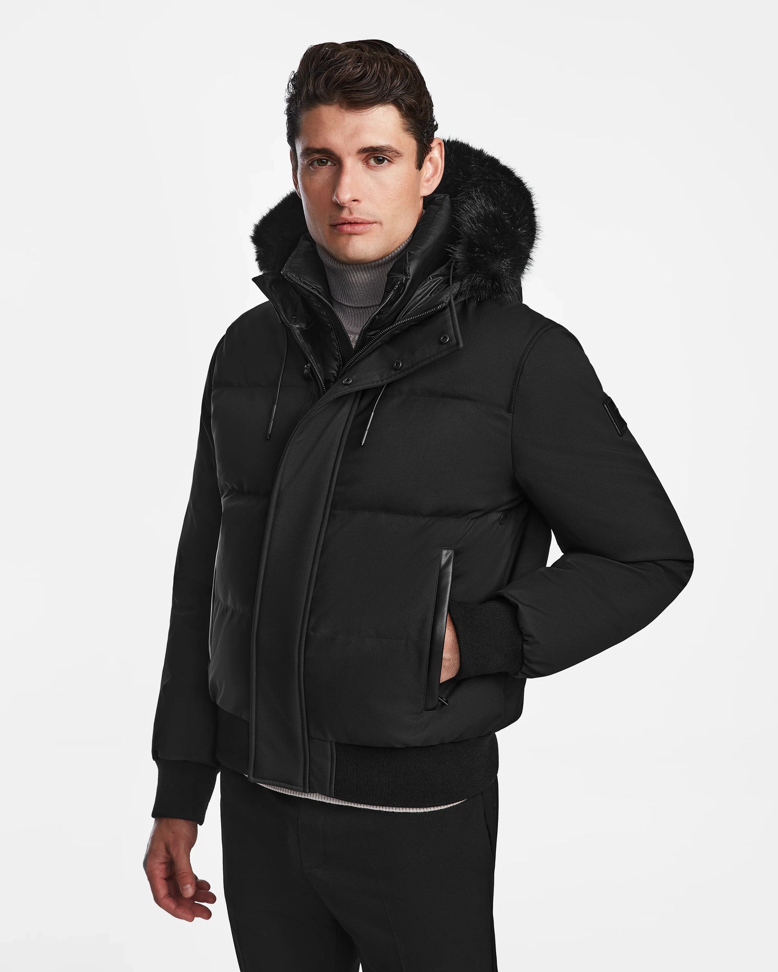 A man models the Rudsak DALLAS - M122501 Black insulated puffer jacket with a fur-lined hood, standing against a plain white background with one hand in his pocket and a neutral expression.