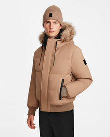 A man models the Rudsak DALLAS - M122501 Sand weather-resistant puffer jacket with fur-lined hood and matching tan knit beanie, paired with black pants, against a plain white background.