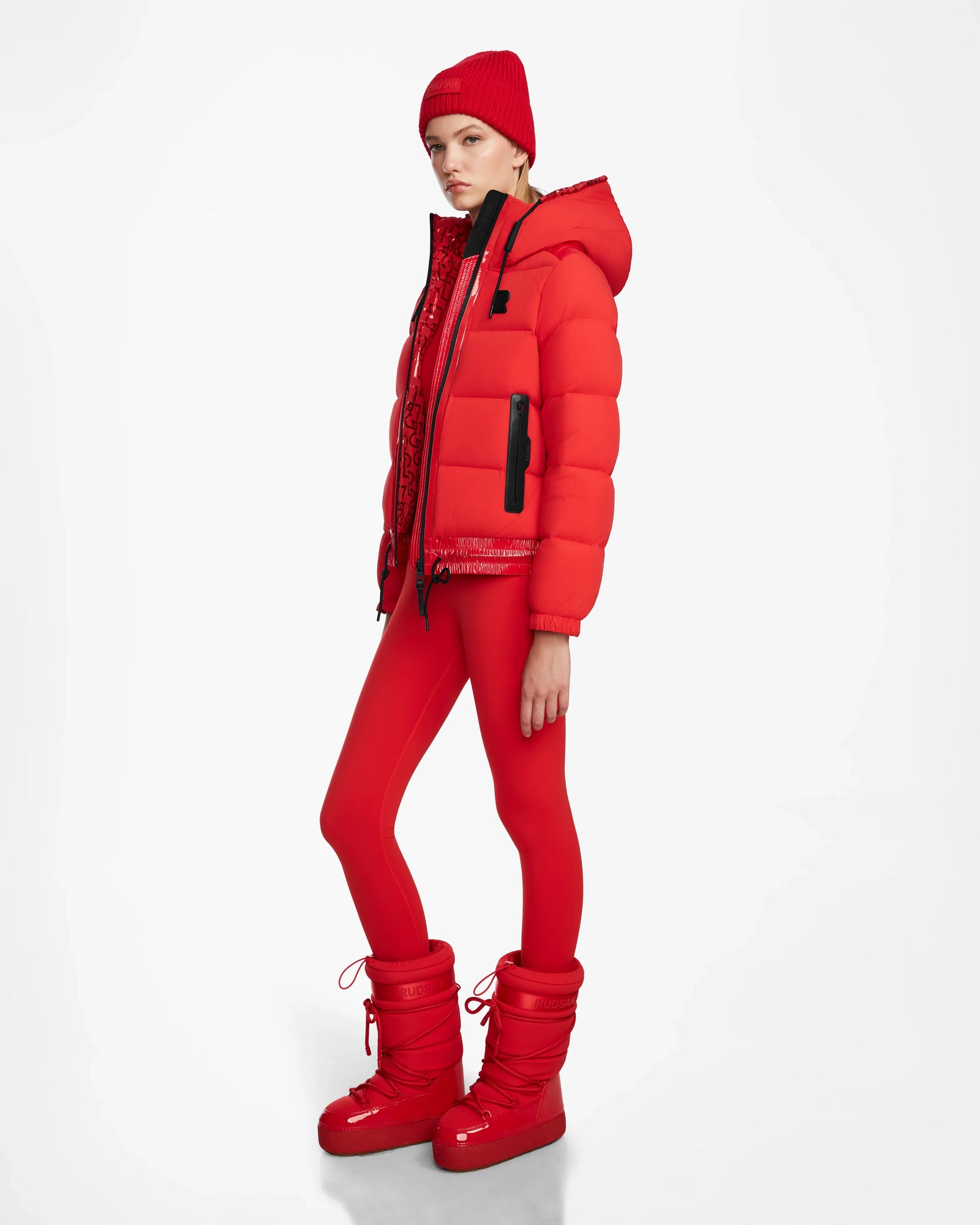 A person stands sideways in a Rudsak DANI M - 8124539 Flame Red winter outfit—faux teddy ski jacket, leggings, beanie, and boots—against a plain white background.