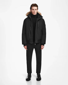 A man faces forward against a white background, wearing the Rudsak DEACON - M123513 Black/Natural bomber-style down puffer with recycled leather trim and RDS-certified down, paired with a black turtleneck, pants, and boots. His hands rest at his sides.