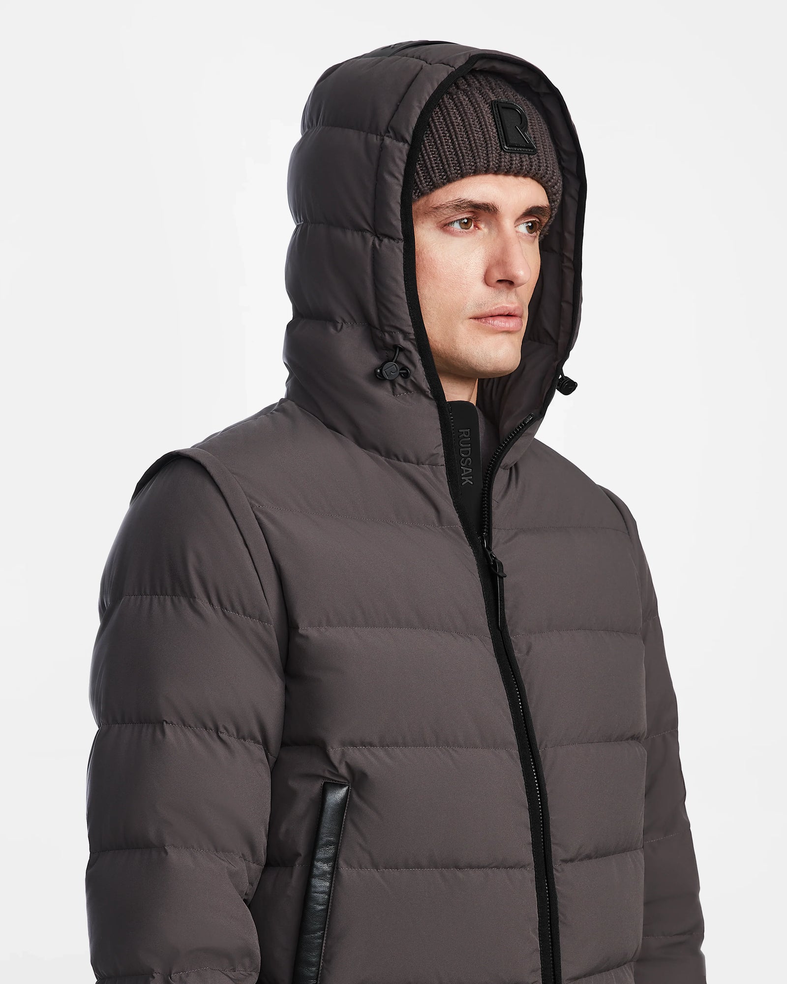 A man wears the Rudsak DEMIAN M-R - M123504 Grey down puffer jacket with a hood up and a ribbed knit beanie, made from sustainable recycled fabric, standing against a plain white background.