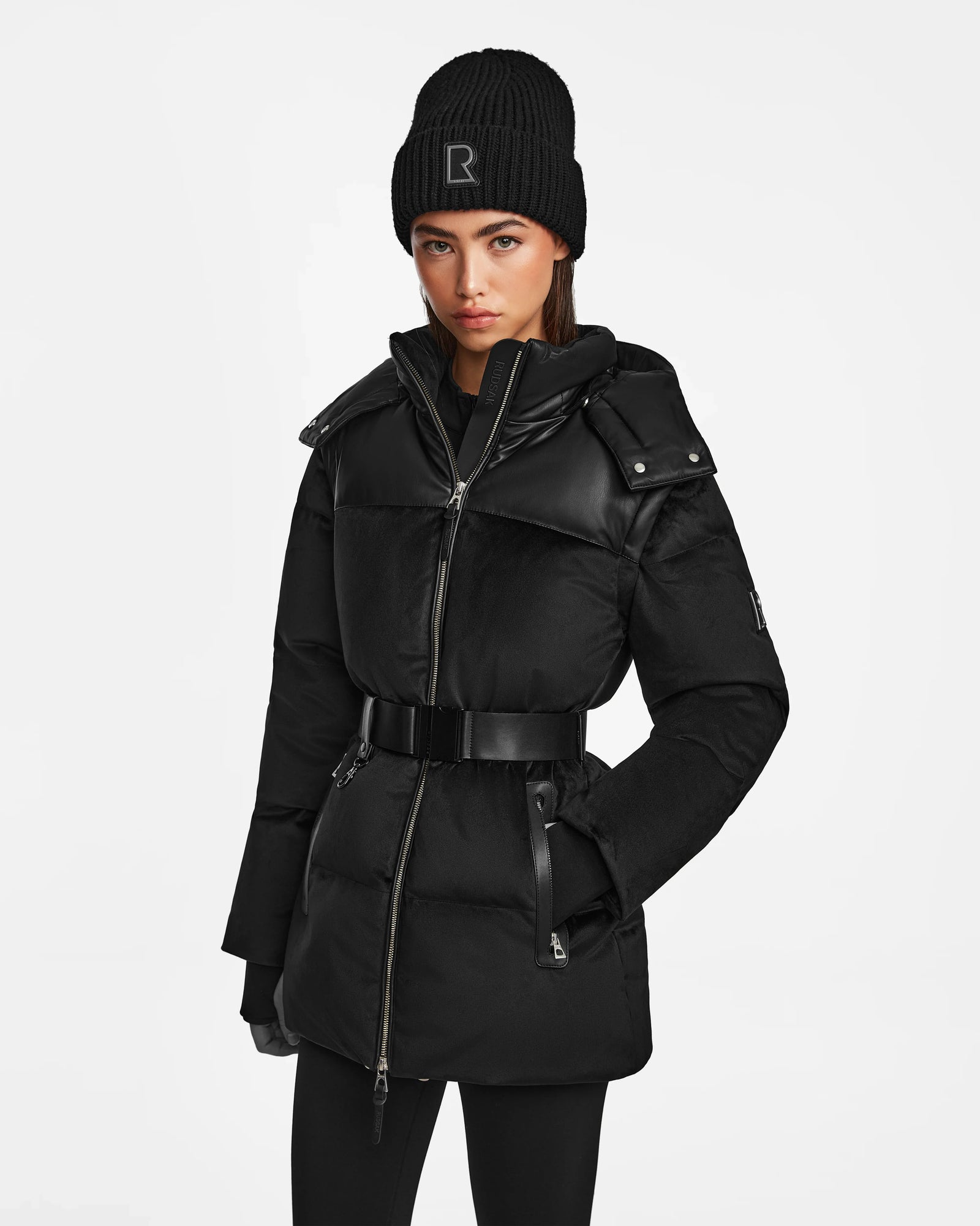 A woman wearing the Rudsak DONIA - L124563 Black convertible winter coat, a black knit hat with an R, black gloves, and black leggings stands against a plain white background.