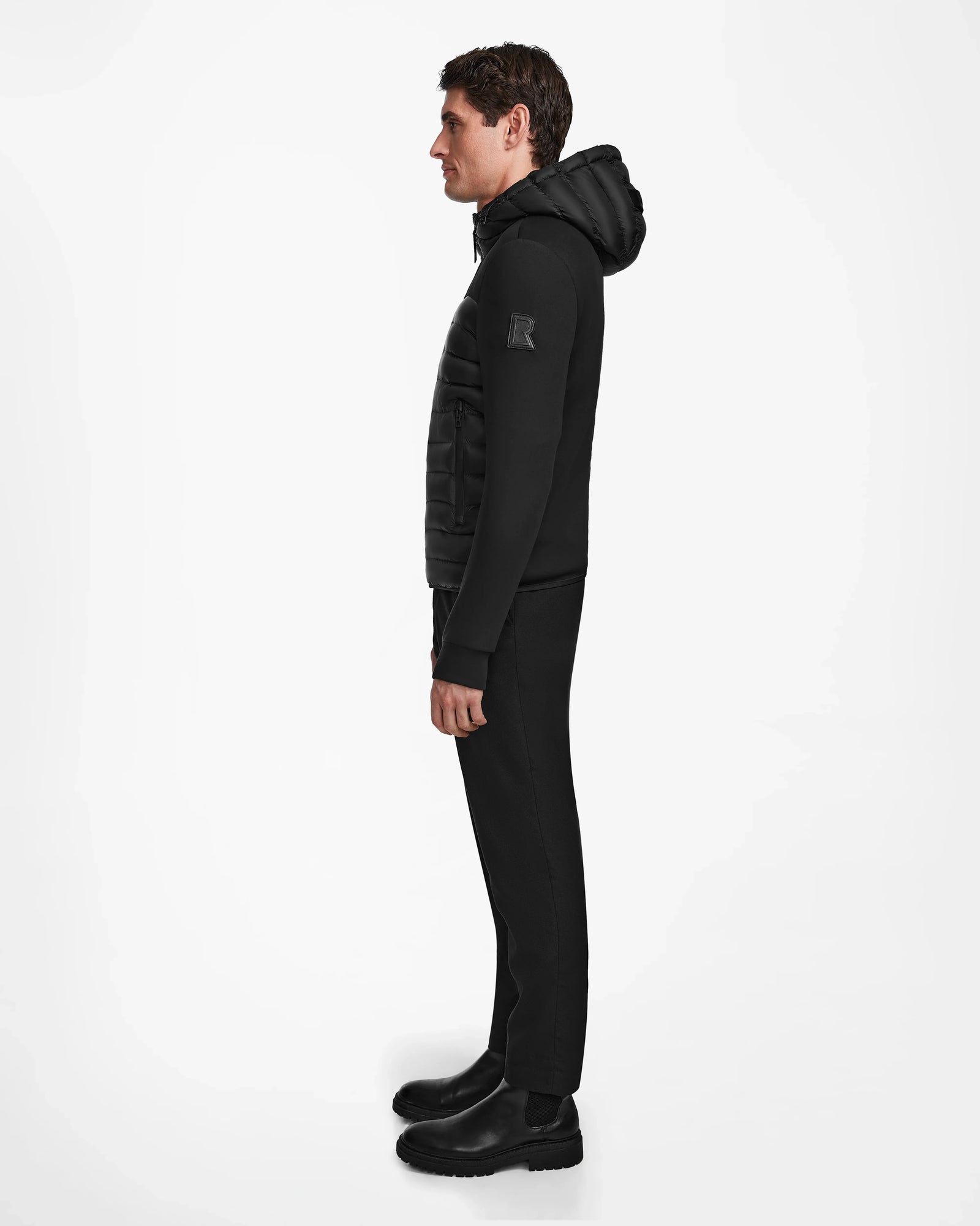 A man stands in profile against a white background, wearing the Rudsak DURIN M - M123515 Black mens hooded jacket with lightweight down insulation, paired with black pants and black boots.