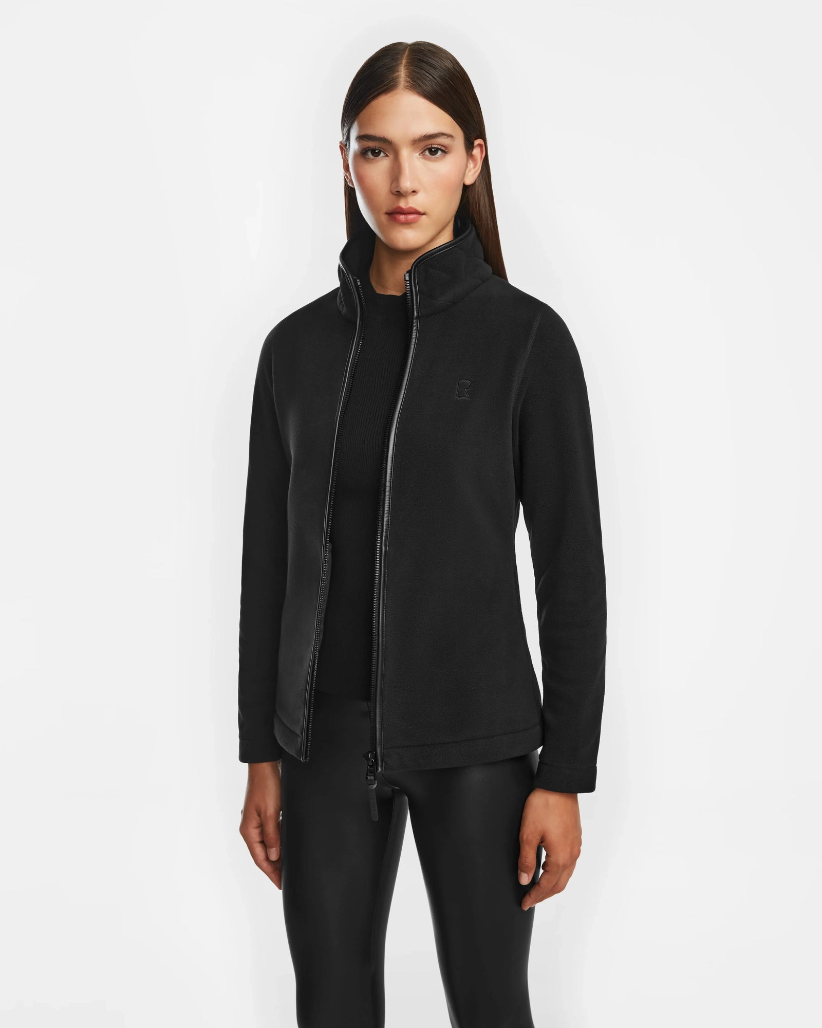 A woman with straight brown hair wears the Rudsak EDITH - 8524617 Black zip-up sweater over a black top and black leggings, standing against a plain white background.