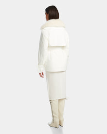 A woman faces away, wearing the Rudsak ELENOR - 8124707 Iceland trench coat with a luxe shearling collar, paired with a white skirt and tall white boots against a plain light gray background.