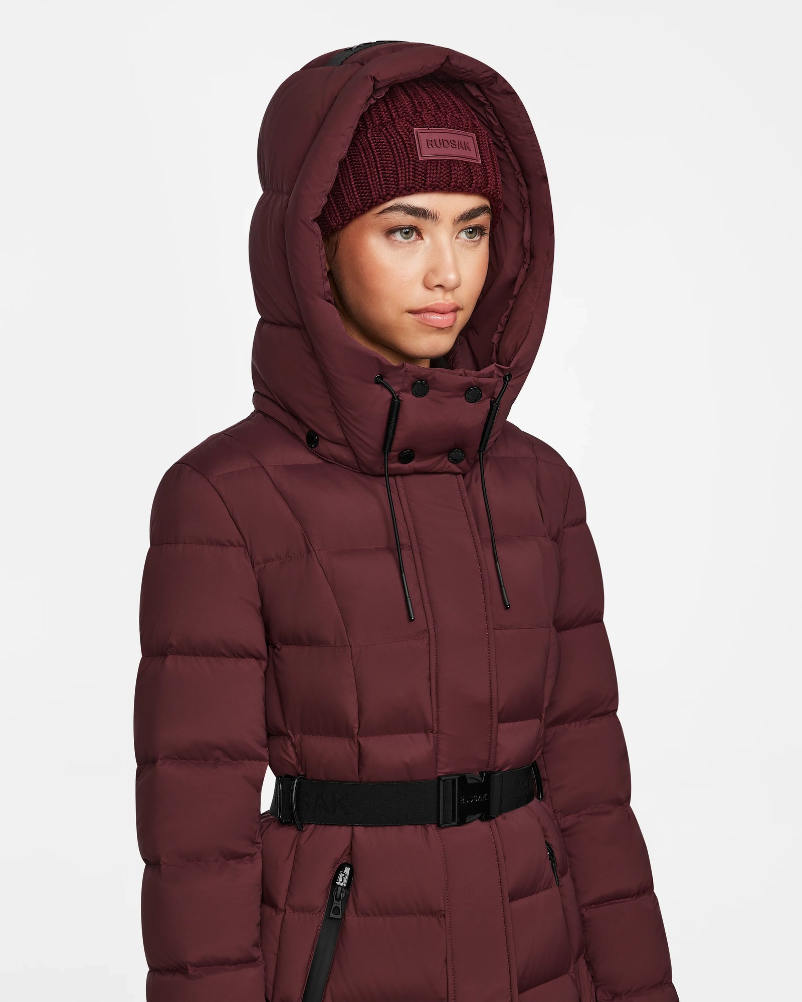 A woman wears the Rudsak FRIDA M - L124524 Wine mid-length down jacket with black accents, a waist belt, and matching knit beanie. The quilted jacket features a removable hood and stands out against the plain white background.
