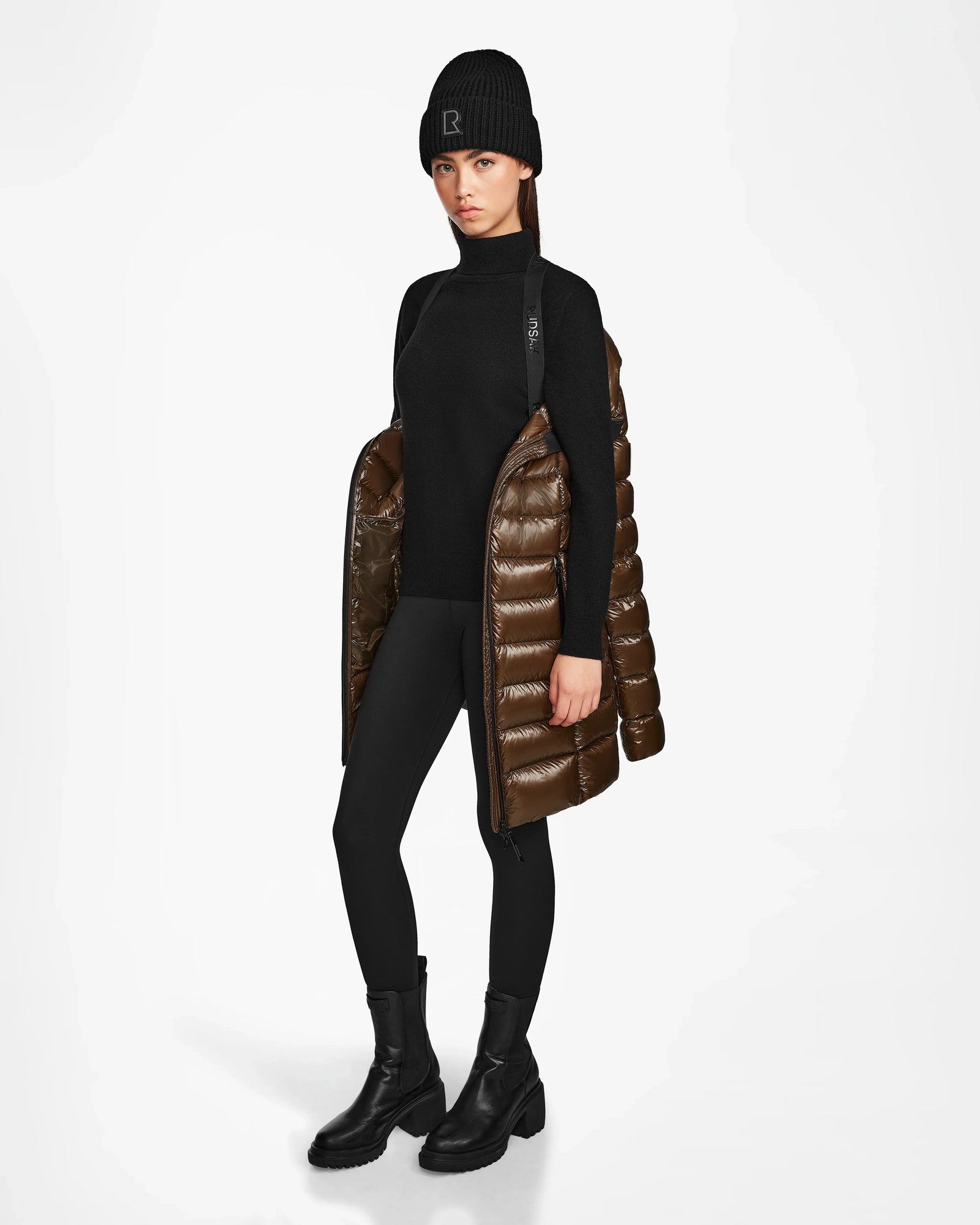 A woman stands against a white background with the Rudsak FRIDA S - L123513 Light Olive women's mid-length down jacket draped over her shoulders, styled with a black knit hat, sweater, leggings, and boots.
