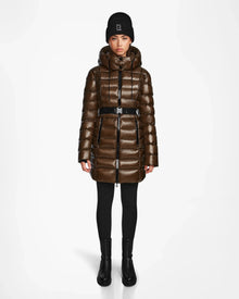 A person stands facing forward wearing the Rudsak FRIDA S - L123513 women’s mid-length down jacket in light olive with a removable hood, black leggings and boots, and a black knit beanie on a plain white background.