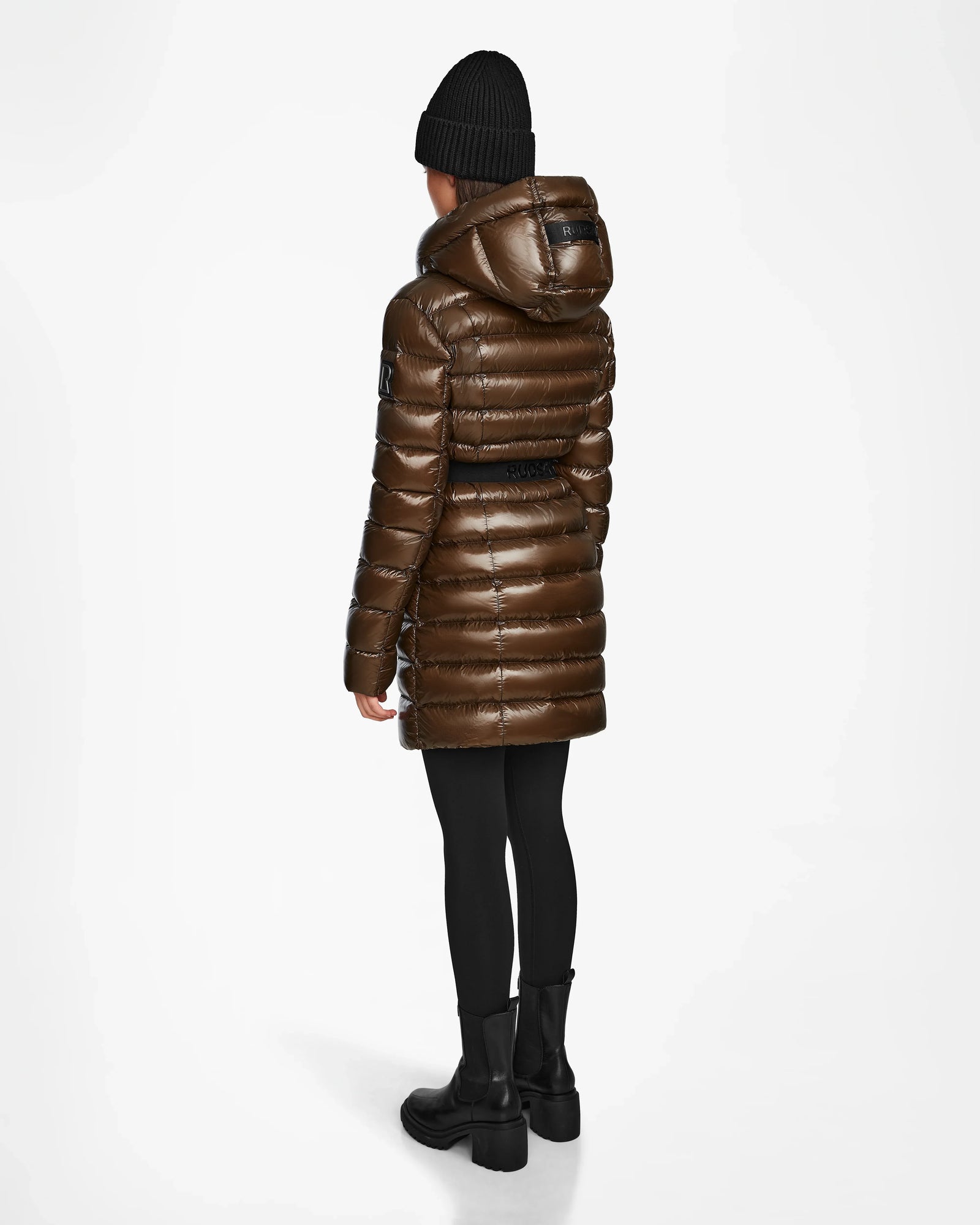 A person stands facing away against a plain white background, wearing the Rudsak FRIDA S - L123513 Light Olive women's shiny mid-length down jacket with removable hood, black leggings, boots, and a black beanie.