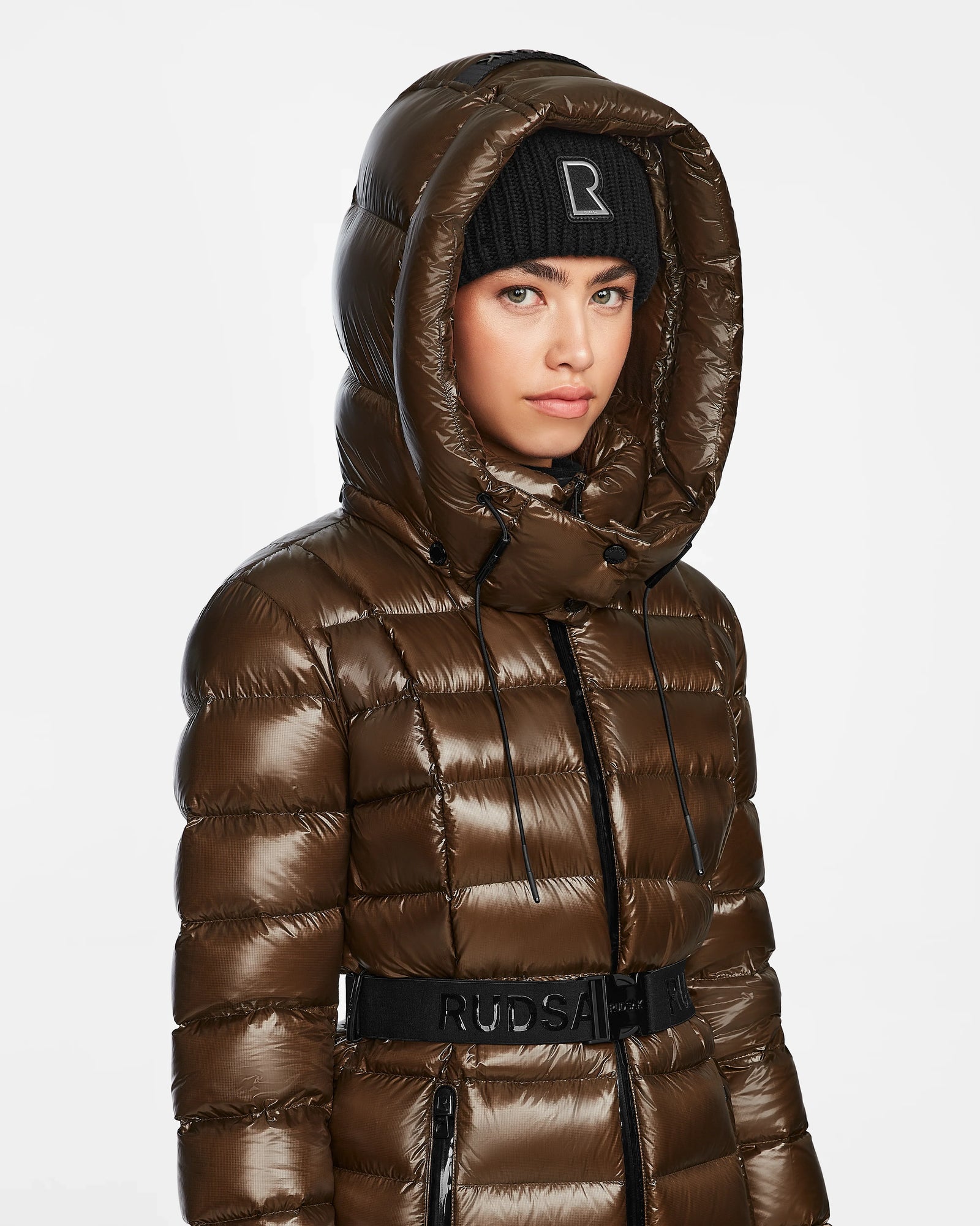 A person wears the Rudsak FRIDA S - L123513 light olive quilted down jacket with a large removable hood and a black knit beanie with a white R logo, standing against a plain white background.