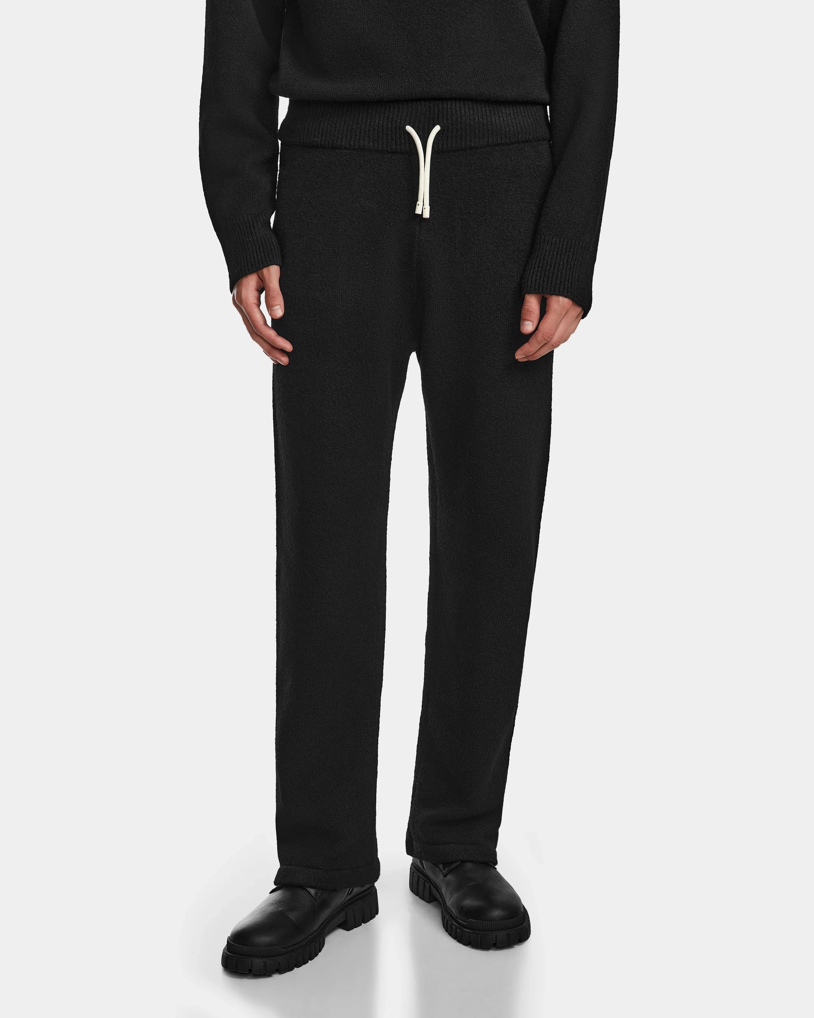 A person wearing Rudsak GEIGER - 9524512 Black M unisex knit lounge pants with a white drawstring, black shoes, and a black long-sleeve top stands against a white background. Only the lower half is shown.