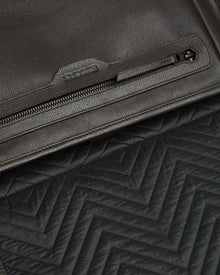 Close-up of the Rudsak GRAYSON - 8324512 Espresso brown leather crossbody bag with a zipper pocket and adjustable strap, shown atop black quilted fabric with a zigzag pattern.