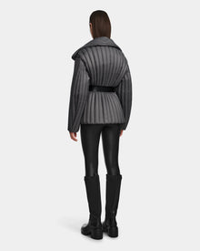 A person faces away, wearing Rudsaks INES T - 8124625 Grey belted quilted jacket with black faux leather pants and knee-high boots against a plain light gray background.
