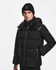 A man wears the Rudsak JACK - M124507 Black quilted jacket with hood and a matching black knit beanie, both featuring the R logo, standing with one hand in his pocket against a plain white background.