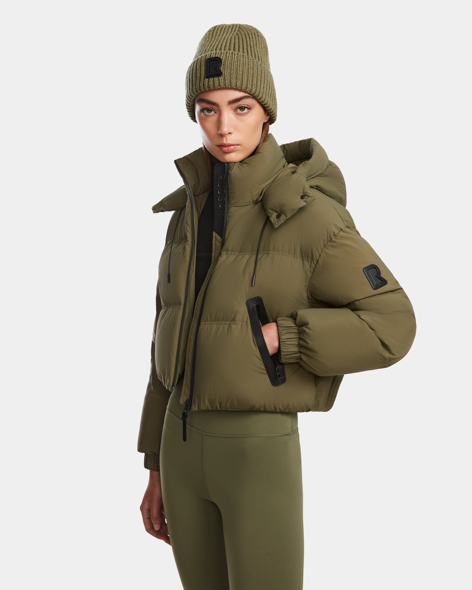 A woman stands against a white background, wearing the Rudsak KARLEE - 8124721 puffer in Loden Green with a matching knit beanie and olive green leggings, looking at the camera with a neutral expression.