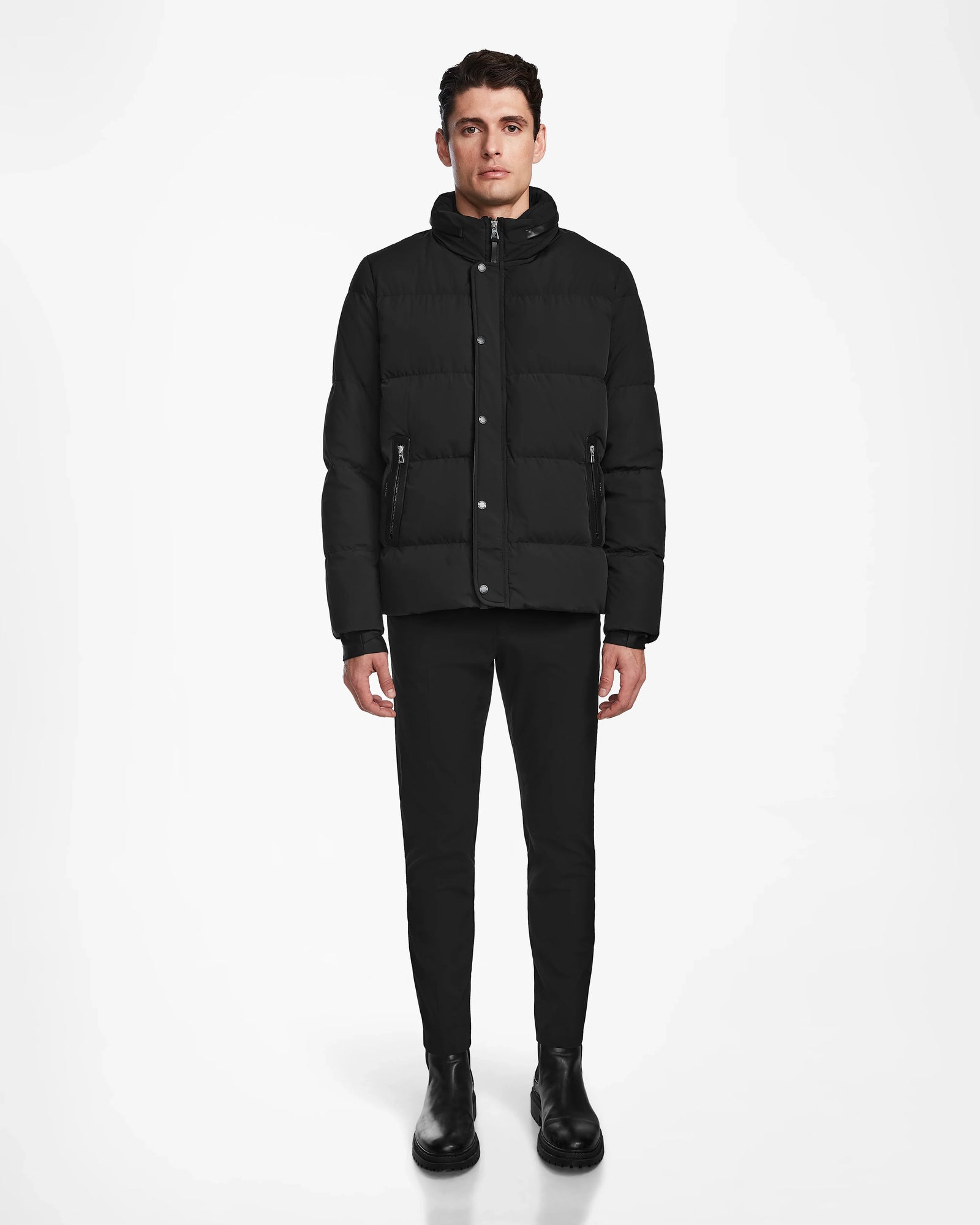 A man stands facing forward against a white background, wearing the Rudsak KARLS - M124505 Black quilted jacket, paired with black pants and boots. His arms are relaxed at his sides.