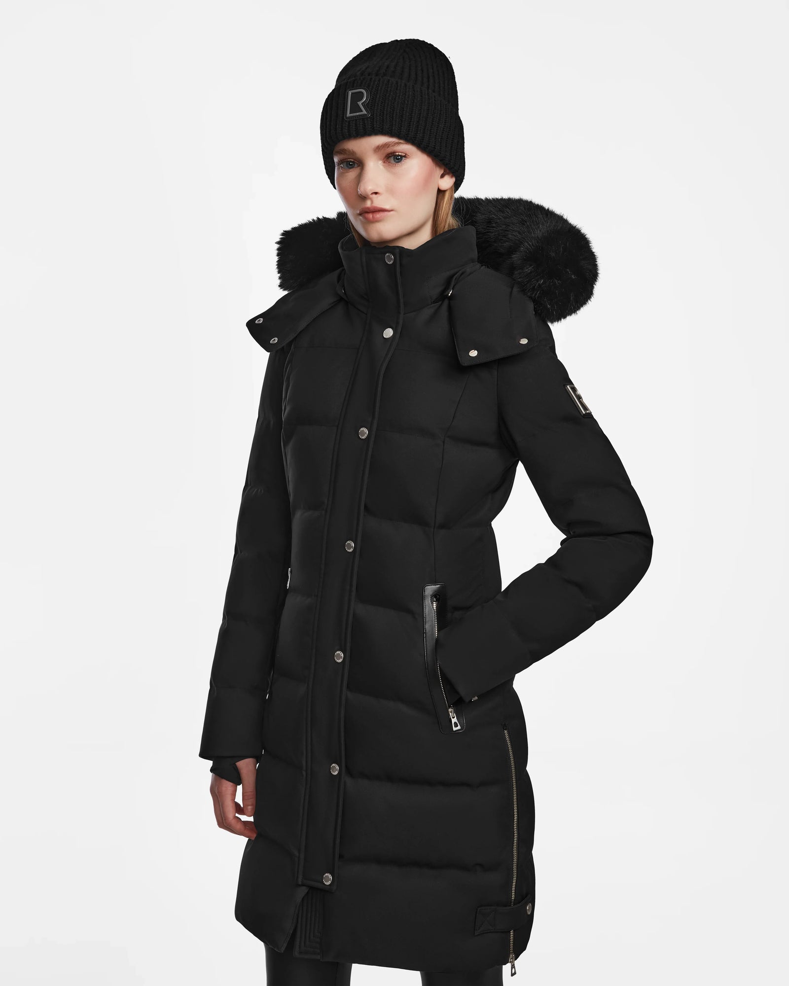 A woman wears the Rudsak KEHLANI - L124540 Black knee-length quilted down coat with a faux fur-trimmed hood and a black knit beanie, standing against a plain white background.