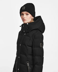 A woman wears the Rudsak KEHLANI - L124540 black quilted down coat with hood and a matching black knit beanie, both featuring an R patch, standing against a plain white background for a true winter essential look.