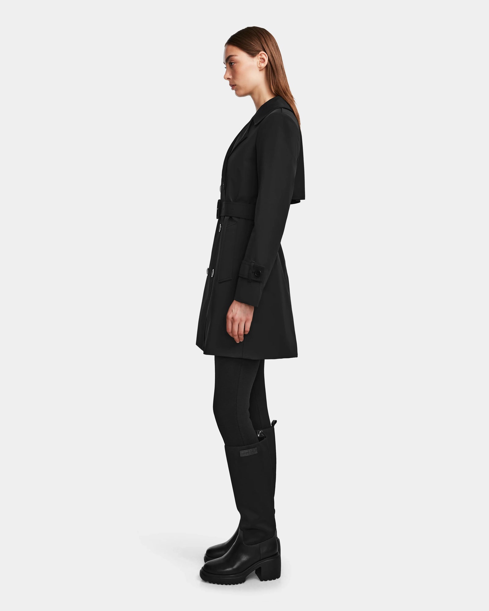 A woman stands in profile wearing the Rudsak KIMBERLY - 8124801 Black trench coat, styled with black leggings and knee-high boots against a plain white background.