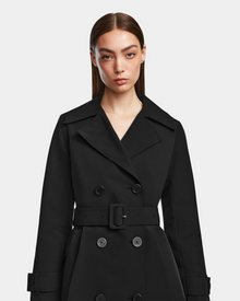 A woman with straight brown hair wears the Rudsak KIMBERLY - 8124801 Black double-breasted womens belted trench coat, standing against a plain light background and looking directly at the camera.
