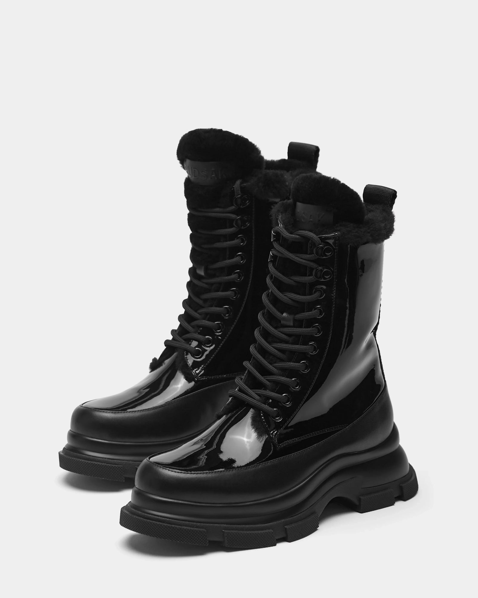 The Rudsak LANA - 8223506 BLACK boots are shiny black mid-calf lace-up boots with chunky soles and soft black shearling lining, shown against a plain white background.