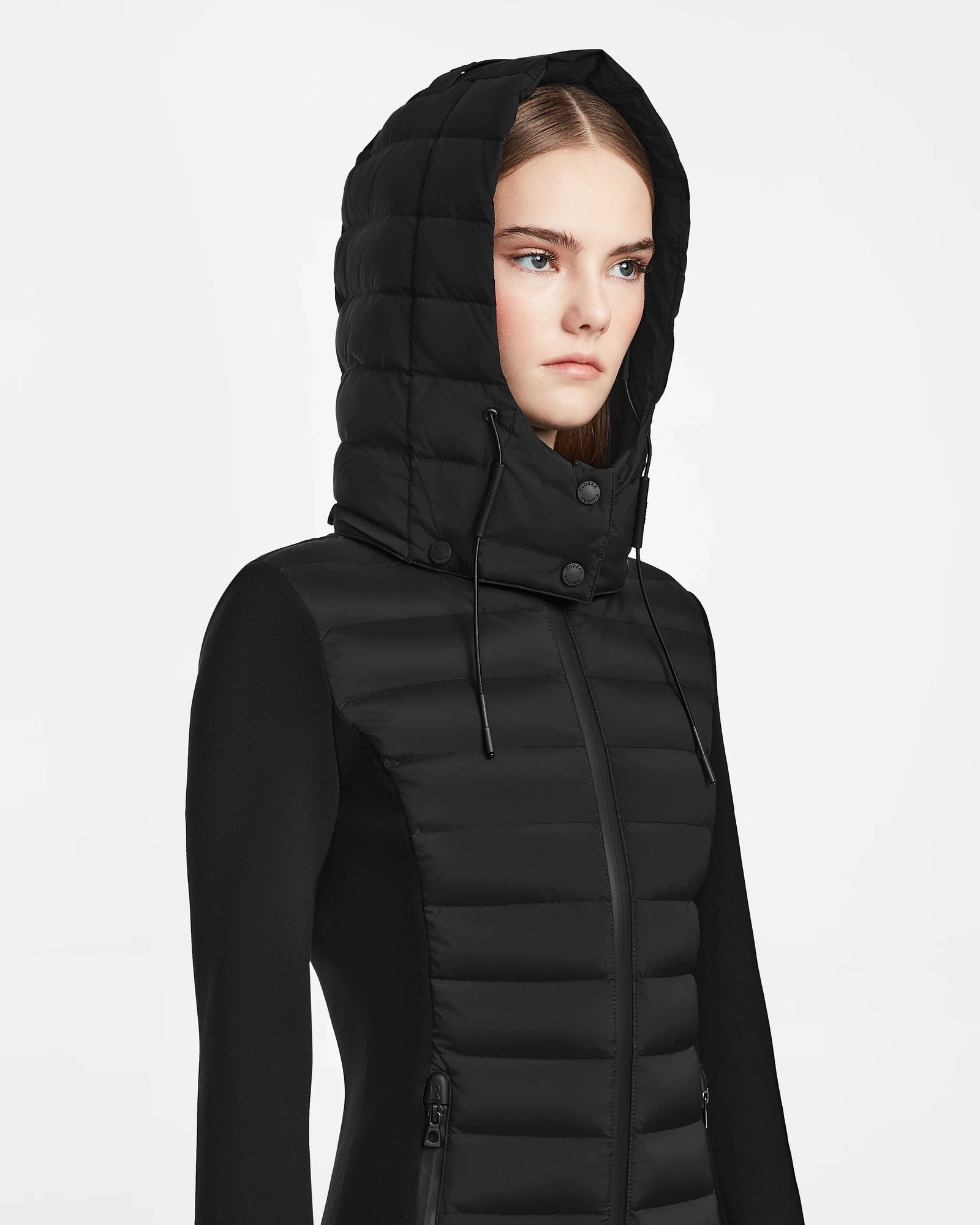 A woman wears the Rudsak MARRY M - L123548 Black women’s puffer jacket with the hood up, looking serious and facing slightly right against a plain white background.