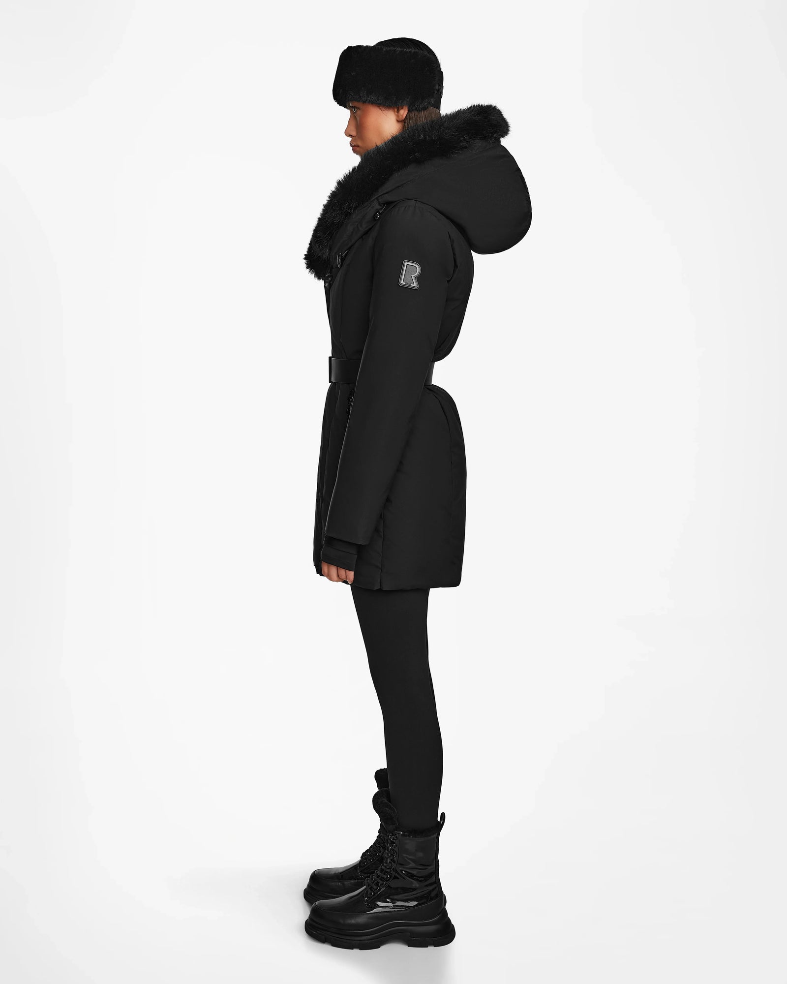 Model in profile, wearing the Rudsak MODA M - 8124555 Black winter coat with a fur collar and hood, styled with black fur hat, leggings, and boots. Sustainable design. Plain white background.