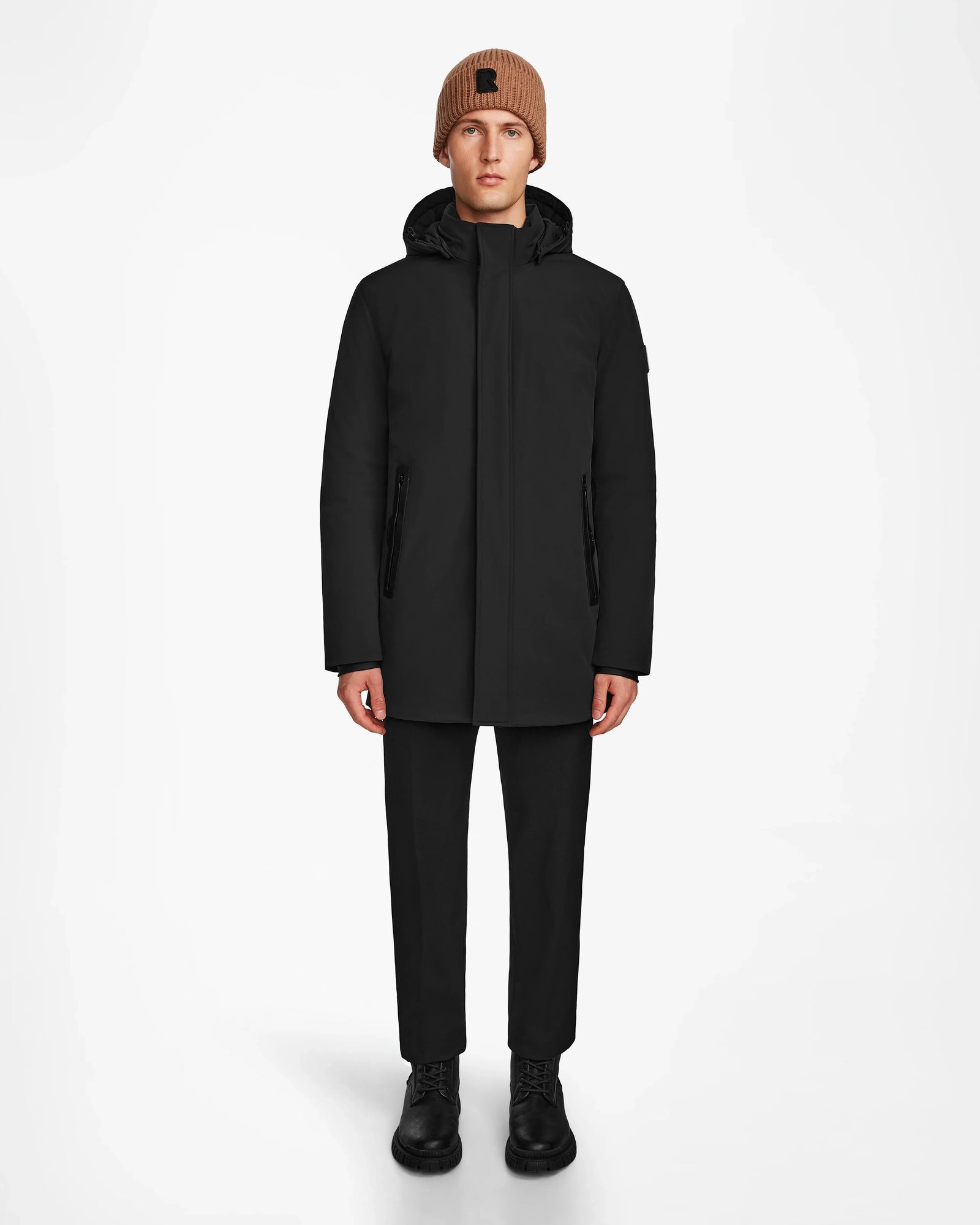 A person faces forward against a plain white background, wearing the Rudsak NATAN - M124519 Black mens parka, paired with black pants, black boots, and a brown knit beanie.