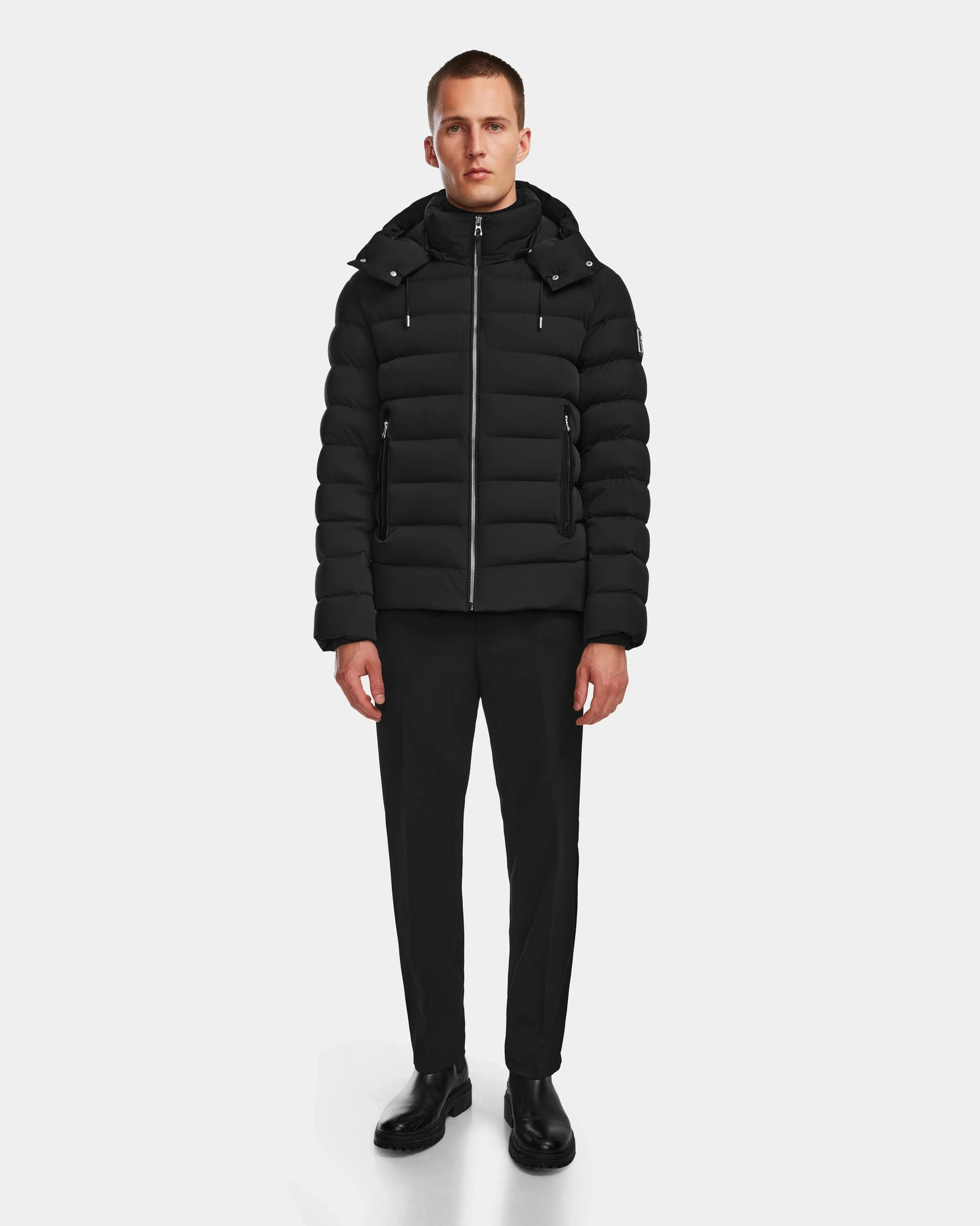 A man stands against a plain background wearing the Rudsak NAVAN - 6124551 Black quilted down puffer jacket with a hood, paired with black pants and shoes, facing forward with a neutral expression.