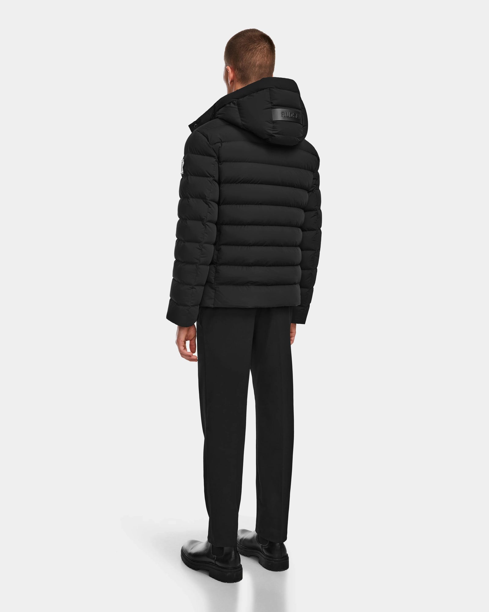 A man faces away wearing the Rudsak NAVAN - 6124551 Black mens quilted down puffer jacket with hood, black pants, and shoes against a plain light background. The jacket features wind- and water-repellent fabric and RDS-certified down fill.