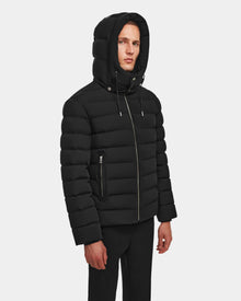 A person wears the Rudsak NAVAN - 6124551 Black men’s quilted down puffer jacket, featuring vertical quilting, zippered side pockets, RDS-certified down, and a wind- and water-repellent finish, against a plain white background.