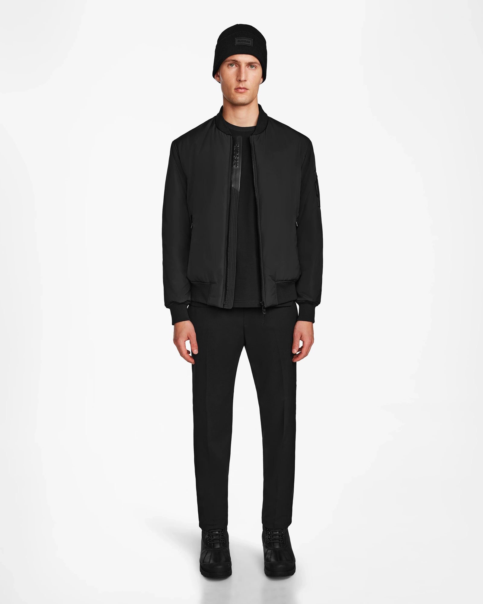 A man faces forward in a Rudsak NEVEN - M124520 Black mens bomber jacket made from recycled materials, styled with a black beanie, shirt, pants, and sneakers against a plain white background.