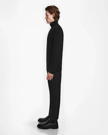 A person with medium-length brown hair stands in profile facing left, wearing Rudsak’s NICOLAS - 6524528 Black cashmere turtleneck and black pants, paired with black boots against a plain white background.