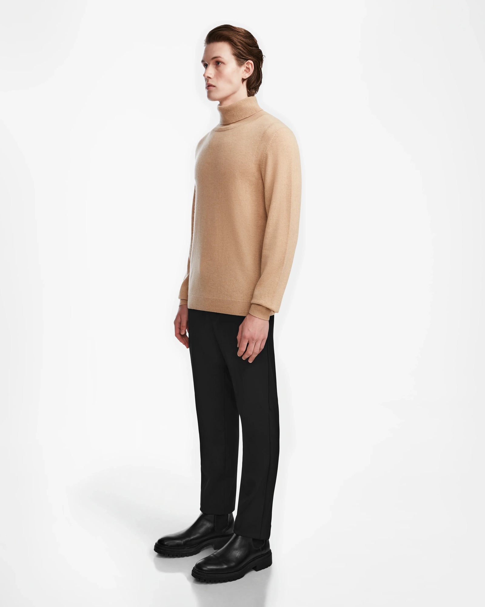 A person stands sideways against a white background, wearing the Rudsak NICOLAS - 6524528 Camel mens sweater, paired with black pants and black shoes.