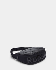 The NIOBE L - 8324503 Black by Rudsak is a quilted black crossbody bag with an adjustable strap and bold shiny black RUDSAK lettering on the front, shown against a plain white background.