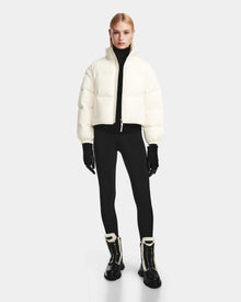 A person stands facing forward wearing the Rudsak OPRAH - 8124708 Iceland womens cropped down puffer jacket with removable sleeves and RDS certified ethical down, paired with a black turtleneck, leggings, gloves, lace-up boots with white fur trim.