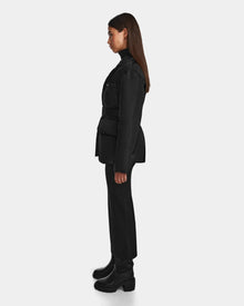 A woman with long brown hair stands in profile facing left, wearing the Rudsak PAOLA - 8124547 Black weather-resistant tailored blazer, paired with black pants, a turtleneck, and chunky-heeled boots against a plain light background.