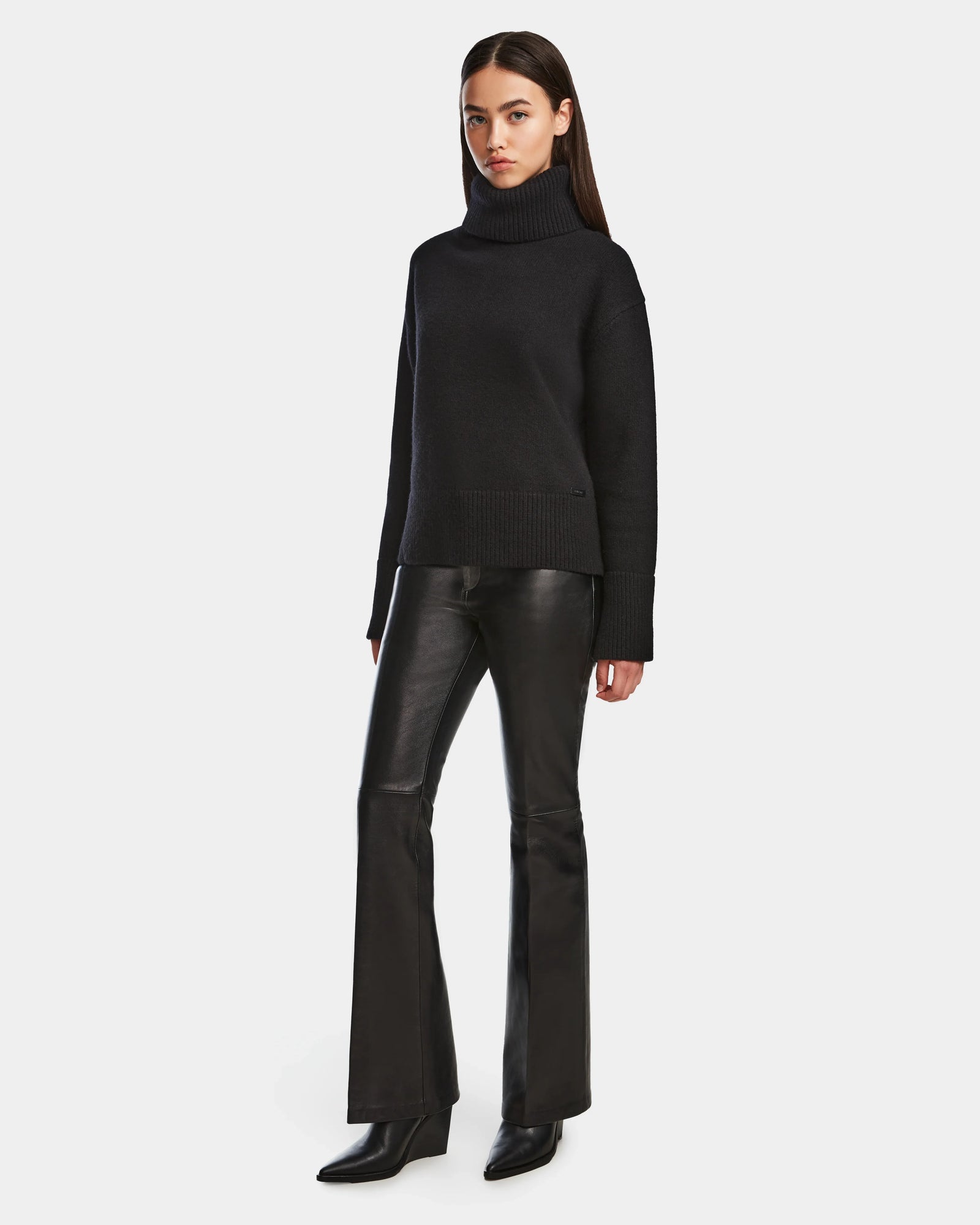 A woman wears the Rudsak PARMI - 8524608 Black turtleneck sweater, paired with black leather flared pants and ankle boots, standing against a plain white background.