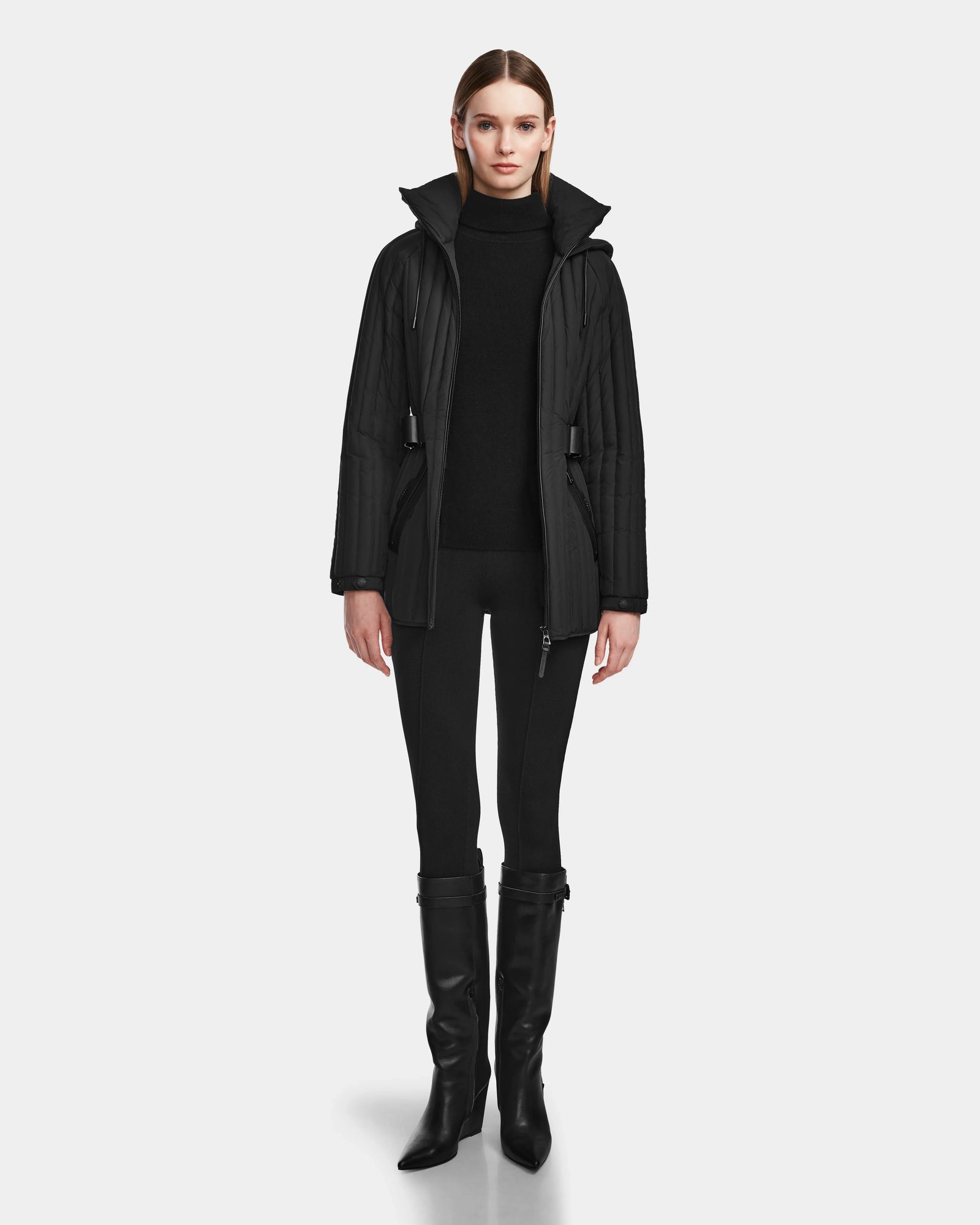 A woman faces forward in a Rudsak PETRAS - 8124725 Black vegan leather quilted jacket with a high collar, black turtleneck, fitted black pants, and knee-high black boots against a plain light background.