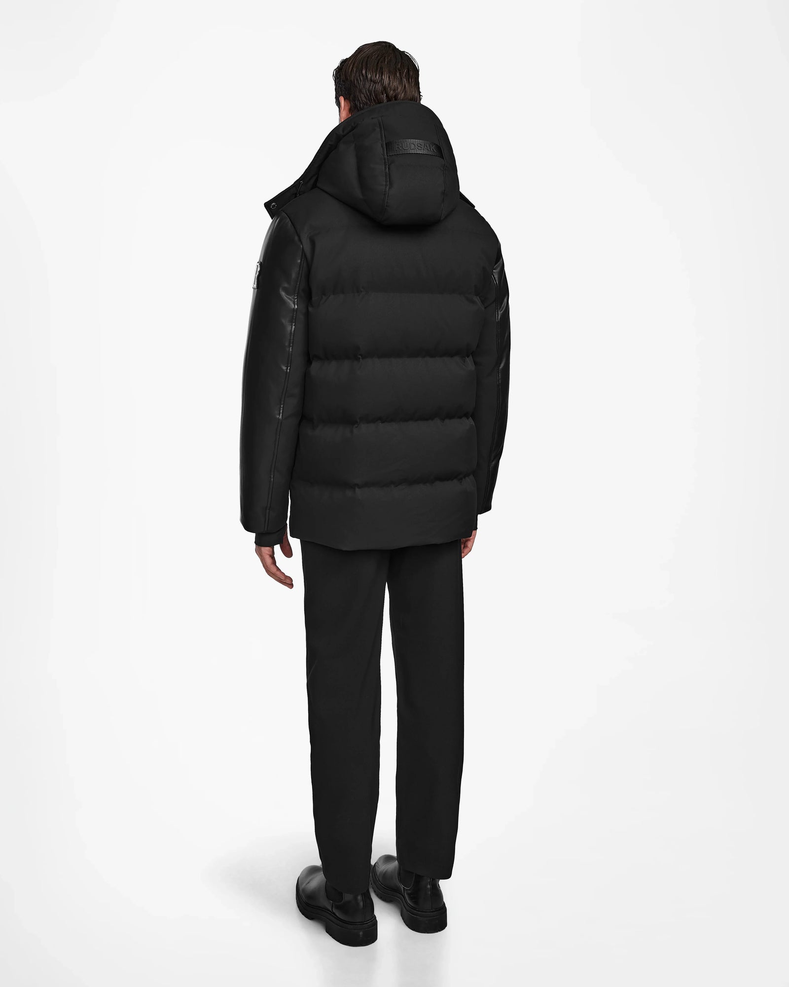 A person stands with their back to the camera, wearing Rudsak's RICKY R - M123506 Black men’s quilted down parka with recycled leather sleeves, black pants, and black shoes against a plain white background.