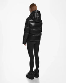 A person with straight, shoulder-length hair models the Rudsak RITA S - L124520 Black weather-resistant puffer jacket, paired with black leggings and sneakers, standing and facing away on a plain white background.