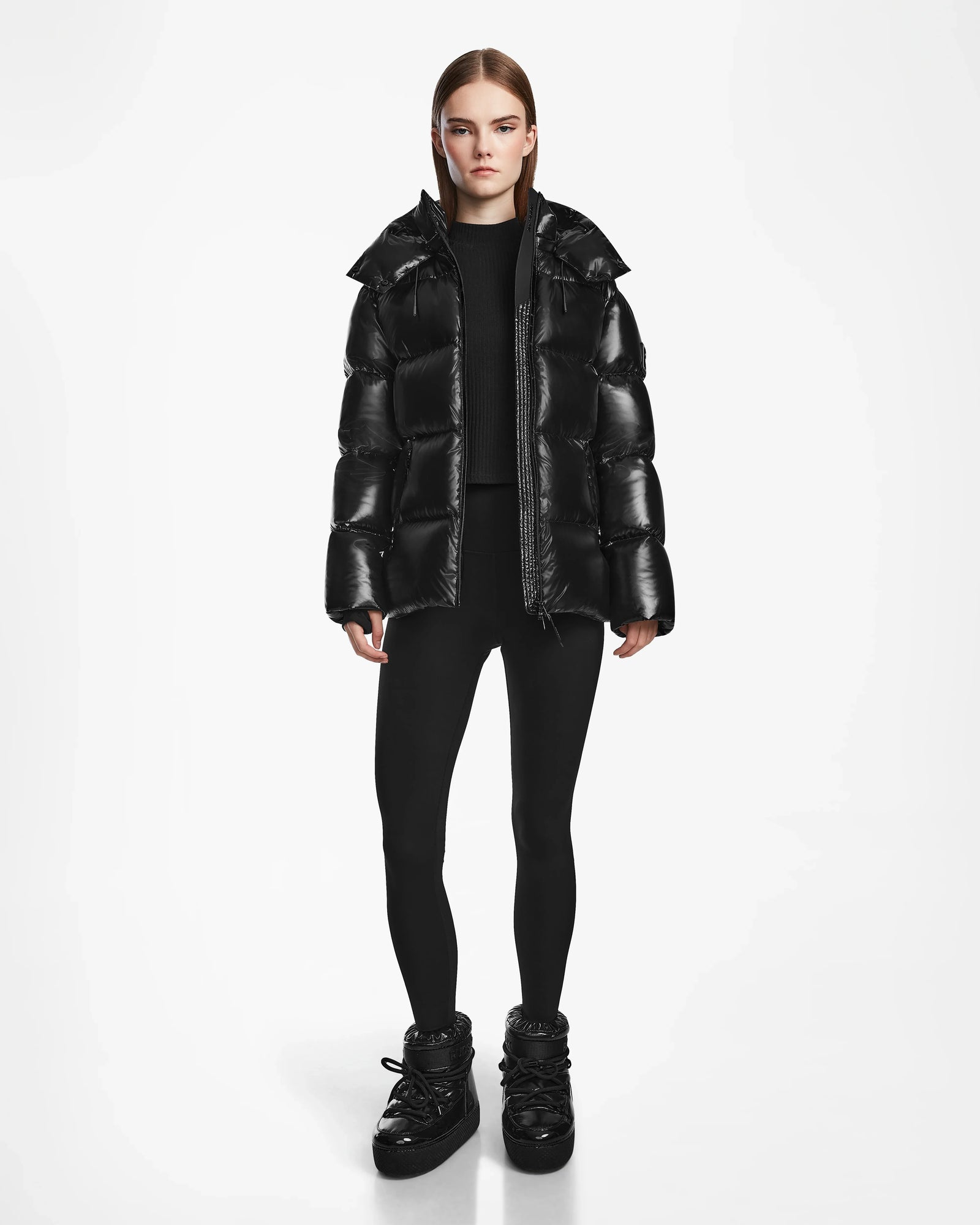 A person wearing the Rudsak RITA S - L124520 Black weather-resistant puffer jacket stands against a plain white background in black leggings and boots, with straight brown hair and a neutral expression, arms relaxed at their sides.