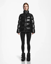 A woman stands facing forward in a shiny black Rudsak RITA S - L124520 weather-resistant puffer jacket, paired with black leggings and boots, against a plain white background.
