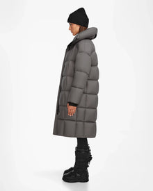 A person wearing the Rudsak SASH - 8124536 Light Grey womens long down puffer coat, black beanie, black pants, and black winter boots stands in profile against a plain white background.