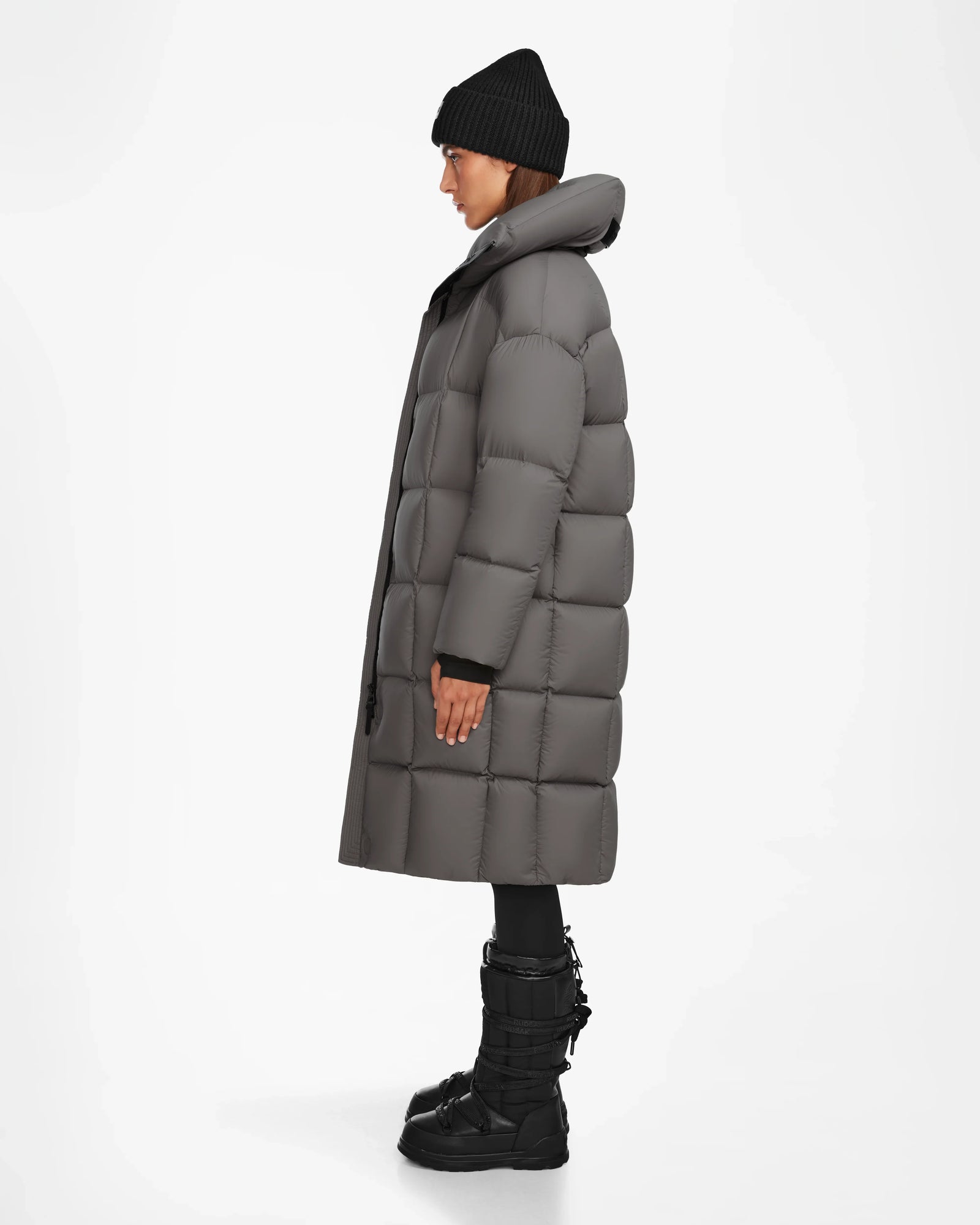 A person wearing the Rudsak SASH - 8124536 Light Grey womens long down puffer coat, black beanie, black pants, and black winter boots stands in profile against a plain white background.