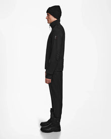 A man wearing the Rudsak TREVOR - 6124519 Black men’s ski jacket with RECCO rescue tech, a black beanie, black pants, and winter boots stands in profile against a plain white background.
